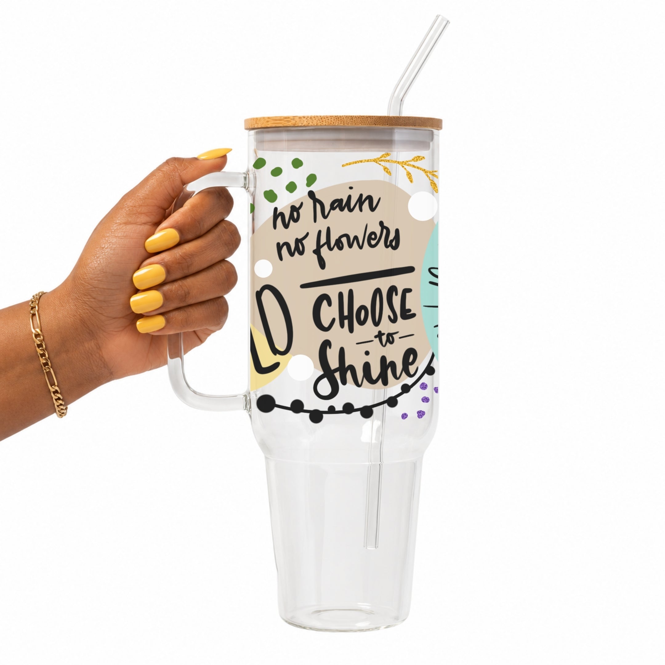 A hand with yellow nails holds a clear handled tumbler with a bamboo lid and clear straw. The tumbler has a printed design with the text 'no rain no flowers' and 'CHOOSE TO SHINE' in black script, with small green, gold, and purple decorative accents.