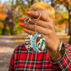 A person outdoors in a red plaid shirt holds up a round clear acrylic keychain with gold hardware. The keychain has a printed design with the text 'Keep Smiling' and 'Tomorrow is a New Day' on a light green background.