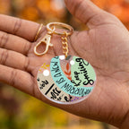A round acrylic keychain with a gold-tone clasp and chain, held in a person's hand. The keychain features a colorful, collage-style design with text including 'Keep Smiling' and 'Tomorrow is a new day' on pastel green, beige, and black sections with small leaf and dot accents.