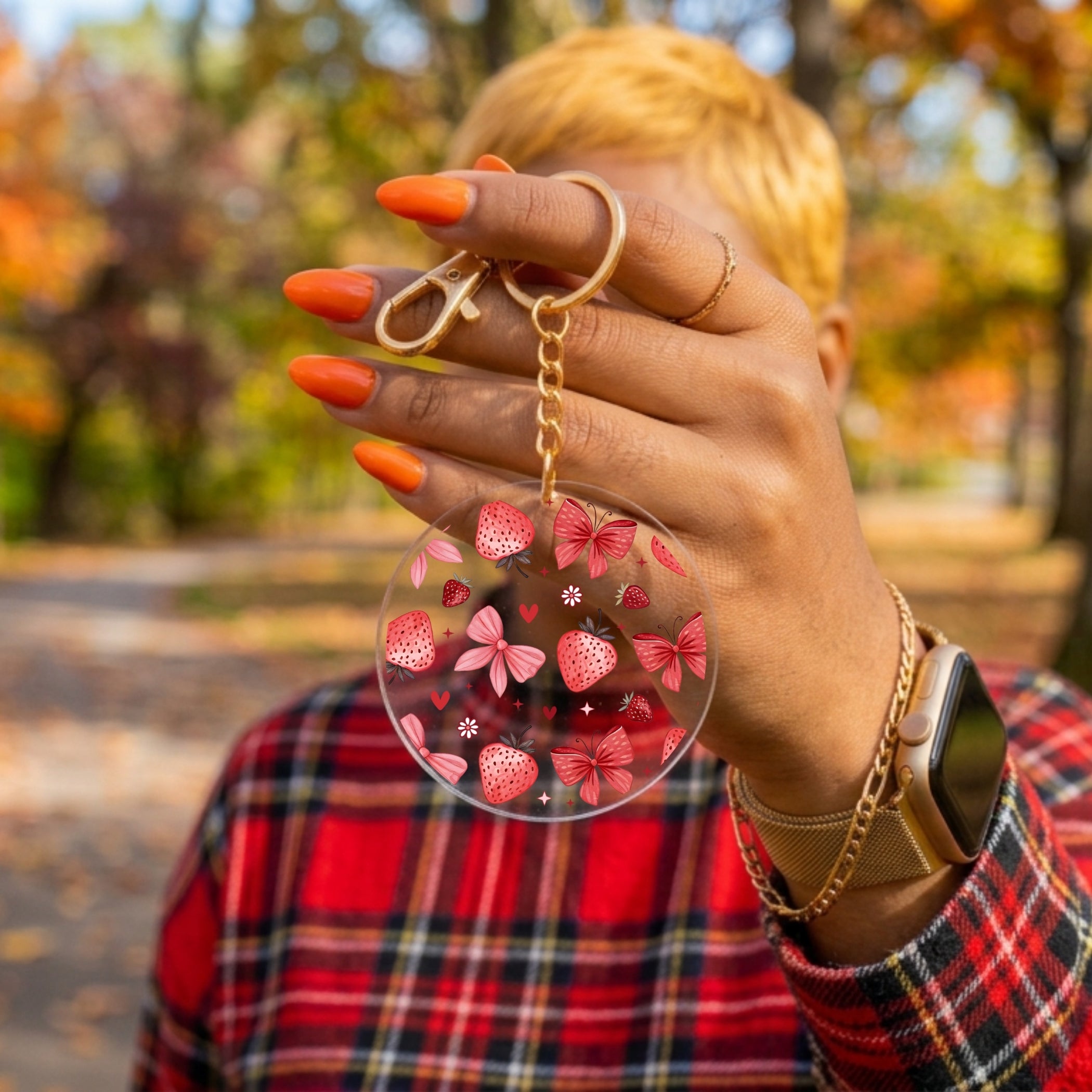 A person outdoors holding a clear round acrylic keychain with pink strawberry and bow patterns and a gold key ring.