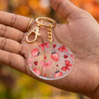 A round acrylic keychain with a strawberry and butterfly bow pattern, held in a person's hand. The keychain features a gold-tone lobster clasp and chain.