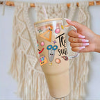 Hand holding a clear 40oz glass tumbler with colorful designs text against a textured beige background