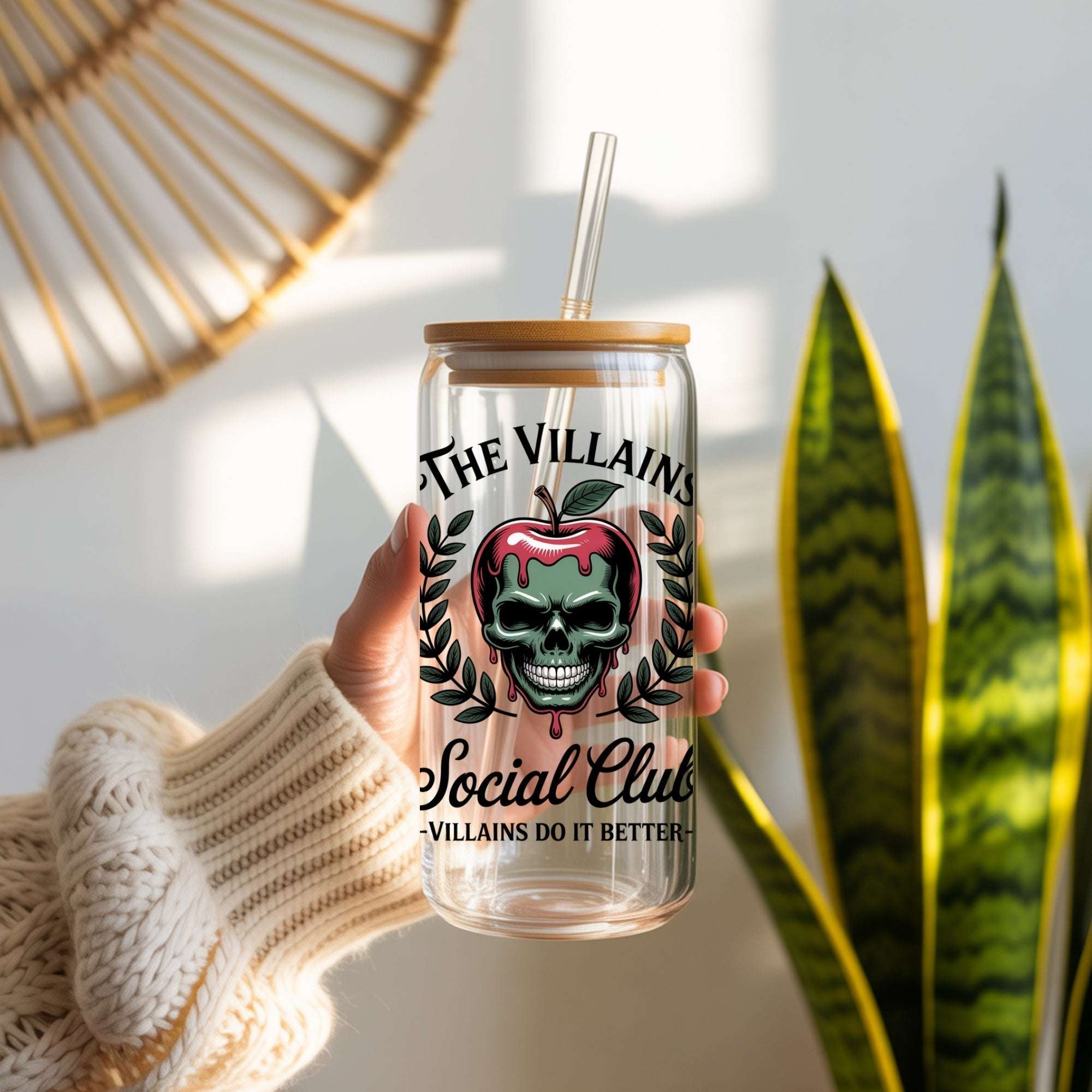 Clear glass tumbler with a skull and apple design, held by a person wearing a beige sweater, with a plant in the background.
