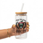 A hand holding a clear glass tumbler with a bamboo lid and clear straw. The tumbler features a graphic of a skull inside a red apple with green leaves, and the text 'THE VILLAINS Social Club' and 'VILLAINS DO IT BETTER'.