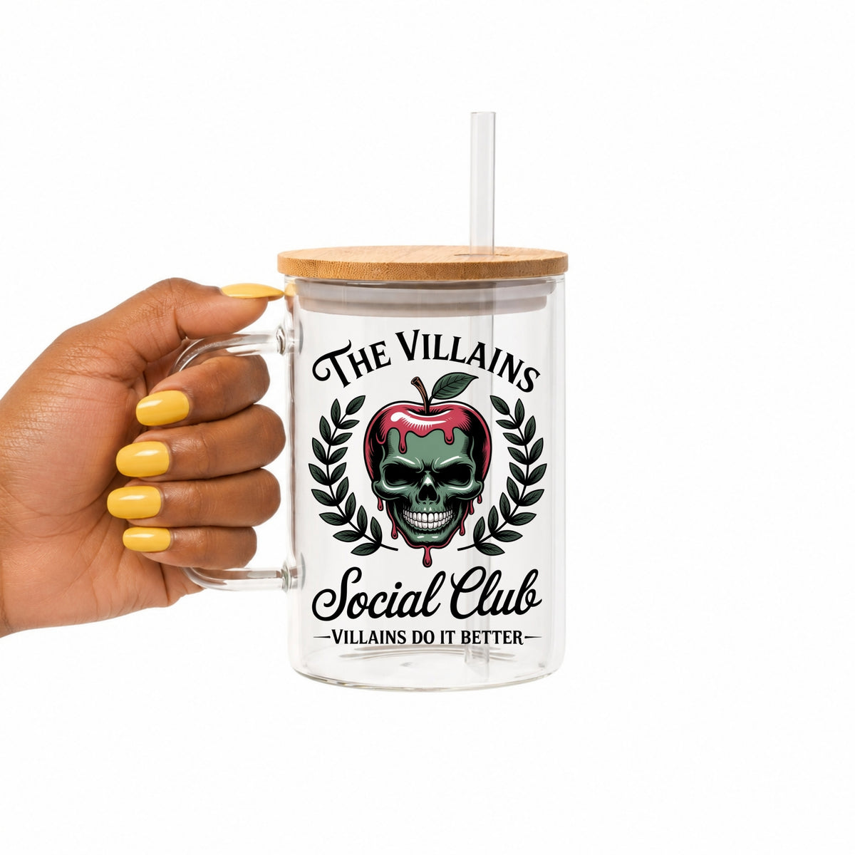 A hand with yellow nails holds a clear glass tumbler with a bamboo lid and straw. The tumbler features a graphic of a skull inside a dripping red apple, framed by laurel leaves, with the text 'THE VILLAINS Social Club' and 'VILLAINS DO IT BETTER'.