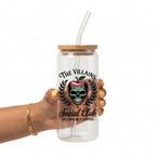 A hand holding a clear glass tumbler with a bamboo lid and clear straw. The tumbler features a graphic of a skull inside a red apple, surrounded by green laurel leaves, with the text 'THE VILLAINS Social Club' and 'VILLAINS DO IT BETTER'.