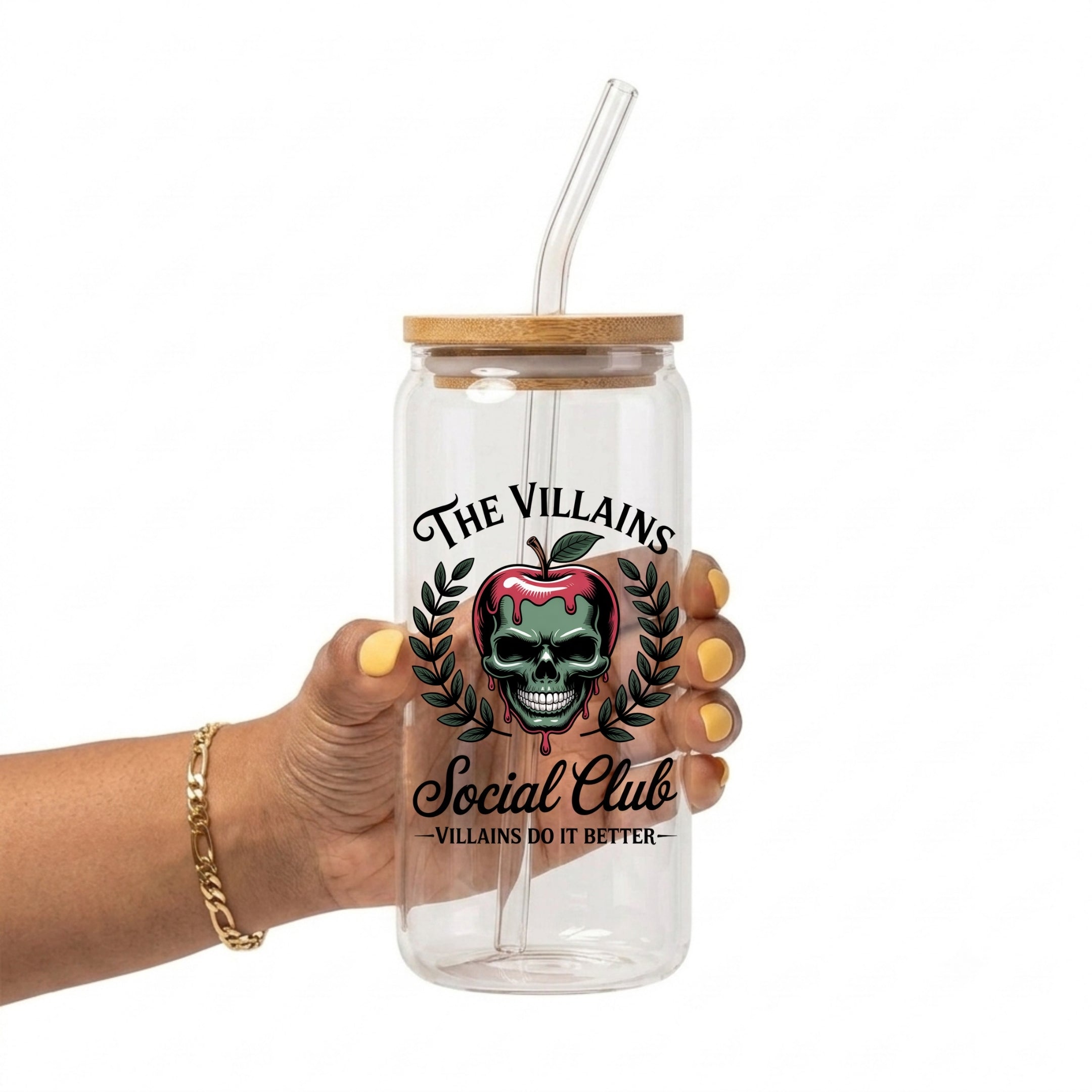 A hand holding a clear glass tumbler with a bamboo lid and clear straw. The tumbler features a graphic of a skull inside a red apple, surrounded by green laurel leaves, with the text 'THE VILLAINS Social Club' and 'VILLAINS DO IT BETTER'.