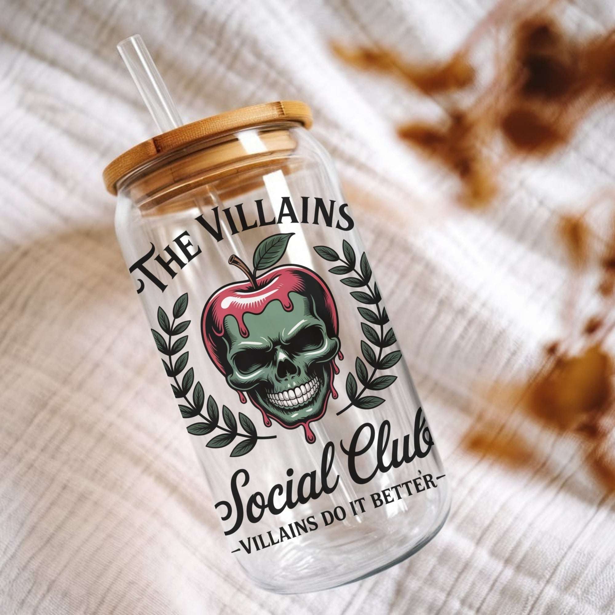 A clear glass tumbler with a bamboo lid and clear straw, printed with a skull inside a dripping red apple, laurel leaves, and the text 'The Villains Social Club -Villains Do It Better-', styled on a textured white fabric background.