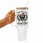 A hand with yellow nails holds a clear handled tumbler with a bamboo lid and straw. The tumbler features a graphic of a skull inside a red apple with black laurel branches and the text 'THE VILLAINS Social Club' and 'VILLAINS DO IT BETTER'.