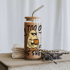 A clear glass tumbler with a bamboo lid and metal straw, filled with a light brown iced coffee or latte. The glass features a cartoon graphic of a ghost wearing sunglasses and holding a mug, with the text 'IM TOO OLD FO THIS SHEET'.