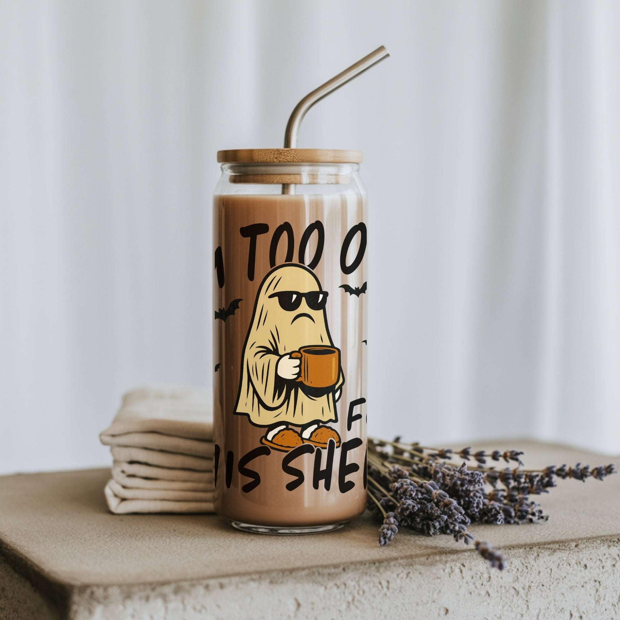 A clear glass tumbler with a bamboo lid and metal straw, filled with a light brown iced coffee or latte. The glass features a cartoon graphic of a ghost wearing sunglasses and holding a mug, with the text 'IM TOO OLD FO THIS SHEET'.
