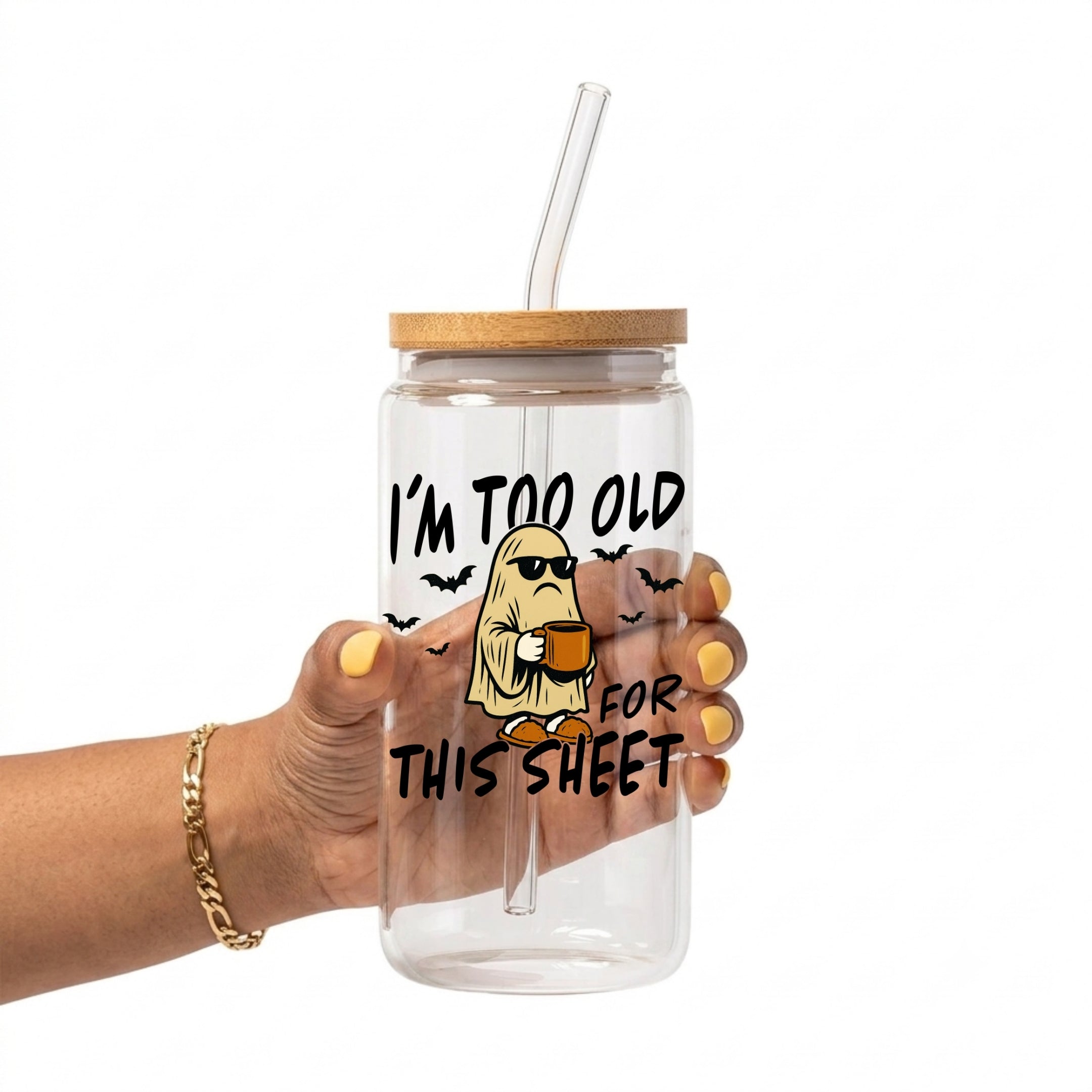 A hand holding a clear glass tumbler with a bamboo lid and clear straw. The tumbler features a cartoon graphic of a ghost wearing sunglasses and holding a mug, with the text 'I'M TOO OLD FOR THIS SHEET' in black.
