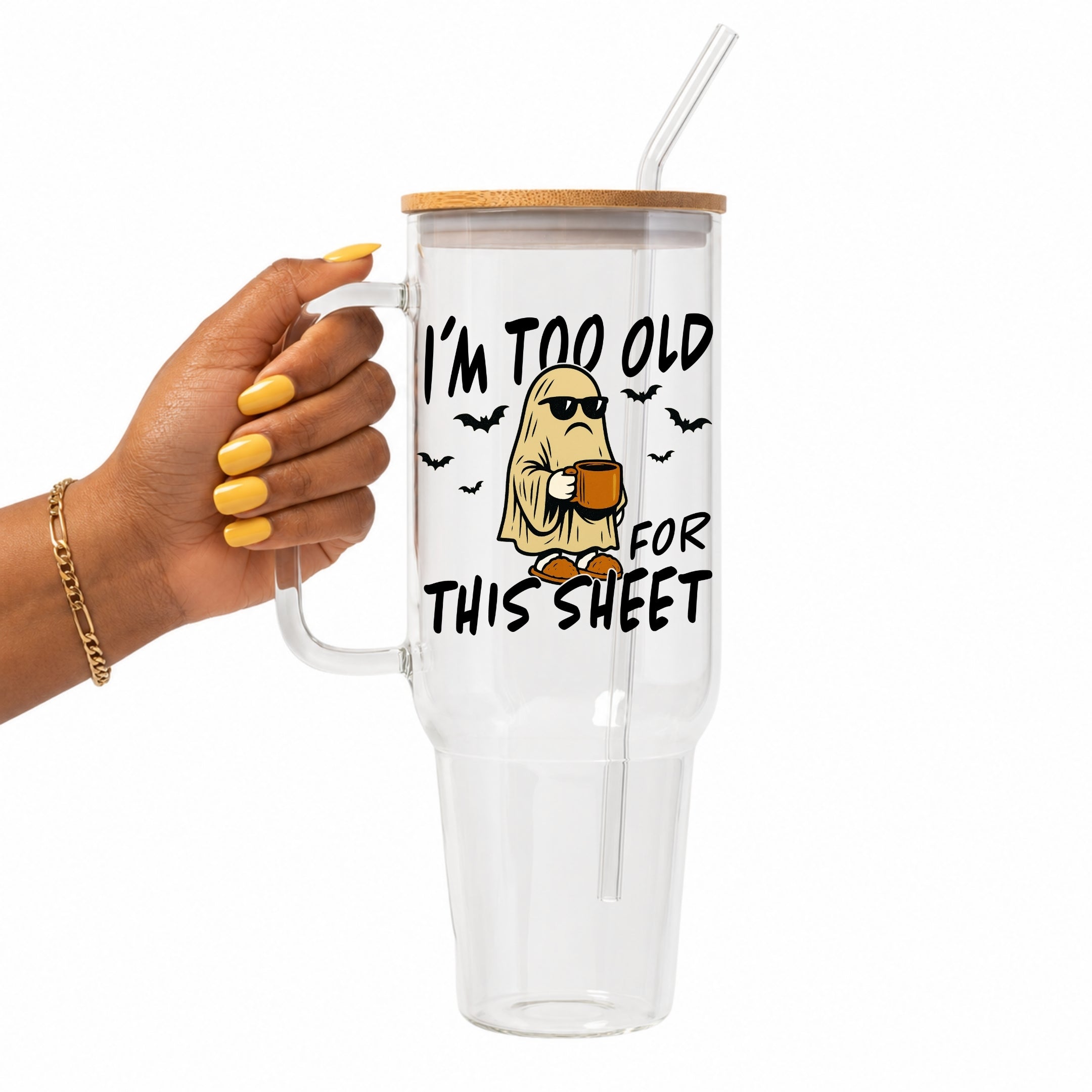 A hand with yellow nails holds a clear glass tumbler with a bamboo lid and straw. The tumbler features a cartoon graphic of a ghost wearing sunglasses and holding a mug, with the text 'I'M TOO OLD FOR THIS SHEET' in black.