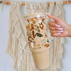 Nik's hand with pink manicured nails holds a clear handled tumbler with a straw, filled with an iced coffee. The tumbler features a beige background decorated with various rustic western-themed illustrations, including a cowboy hat, boots, horseshoe, cactus, and stars.