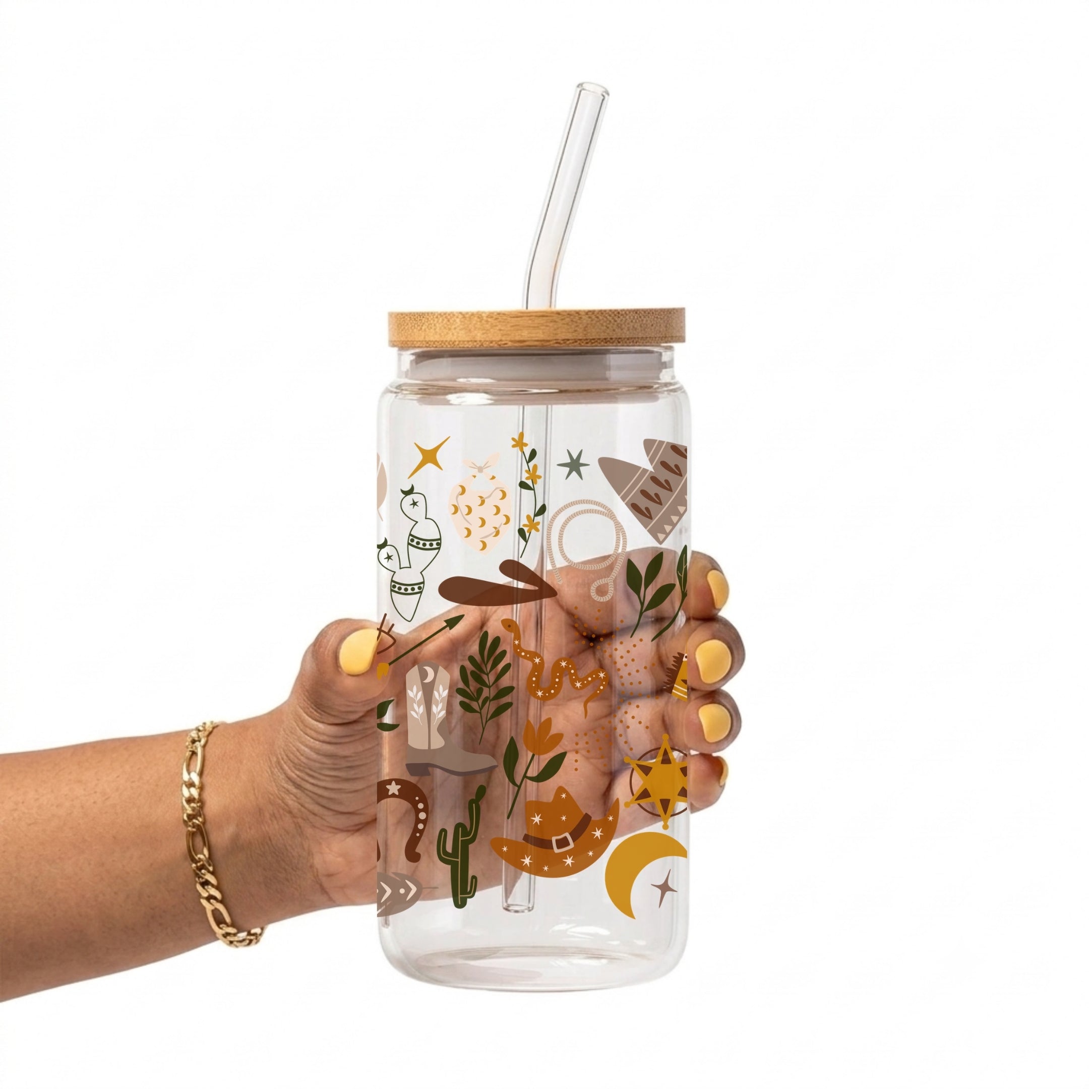 A hand holding a clear glass tumbler with a bamboo lid and clear straw, decorated with a whimsical desert-themed illustration in earthy tones of brown, tan, green, and orange.