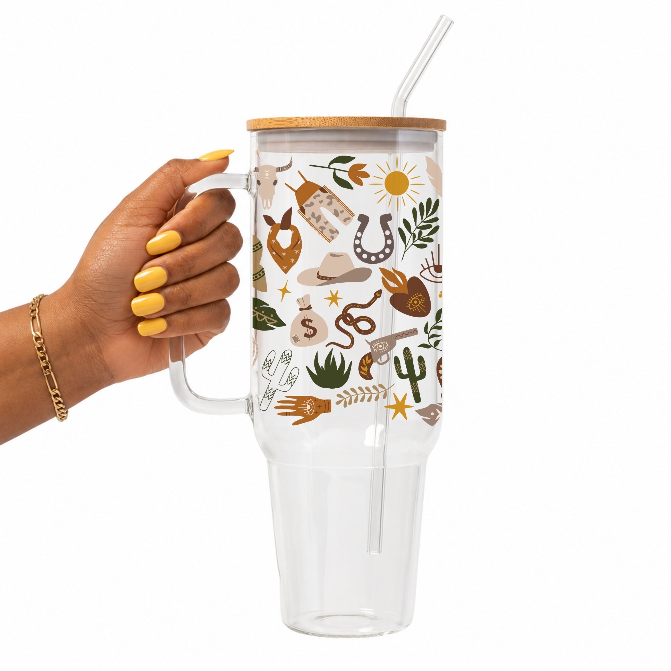 A hand with yellow nail polish holds a clear handled tumbler with a bamboo lid and clear straw. The tumbler is decorated with a western-themed pattern featuring a horse skull, horseshoe, cowboy hat, boots, cactus, and desert plants.