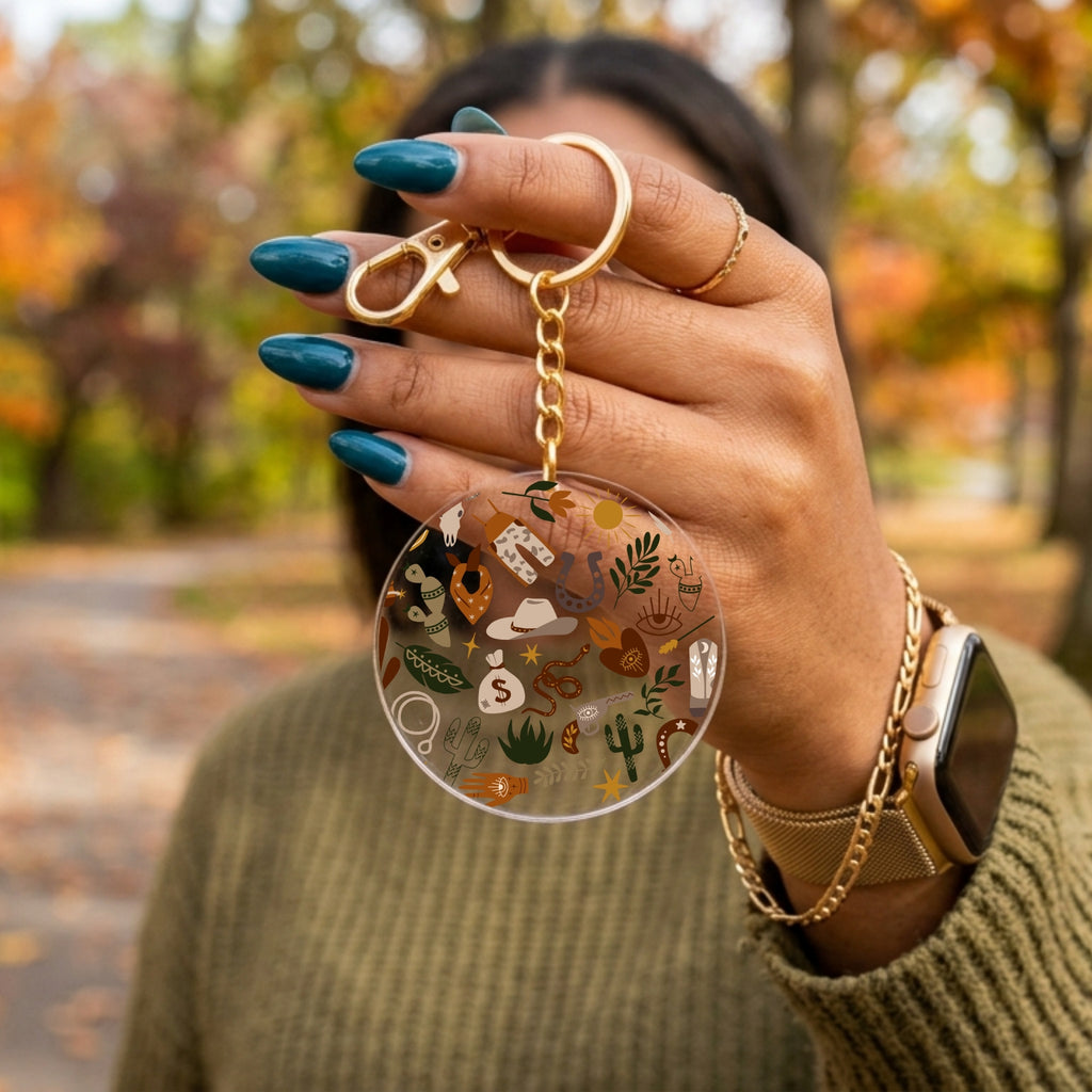 A person outdoors holds up a round clear acrylic keychain with a gold clasp and chain. The keychain features a printed design of various desert and southwestern-themed illustrations, including a cowboy hat, cactus, and sunflower, set against a blurred autumn park background.