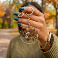A person outdoors holds up a round clear acrylic keychain with a gold clasp and chain. The keychain features a printed design of various desert and southwestern-themed illustrations, including a cowboy hat, cactus, and sunflower, set against a blurred autumn park background.