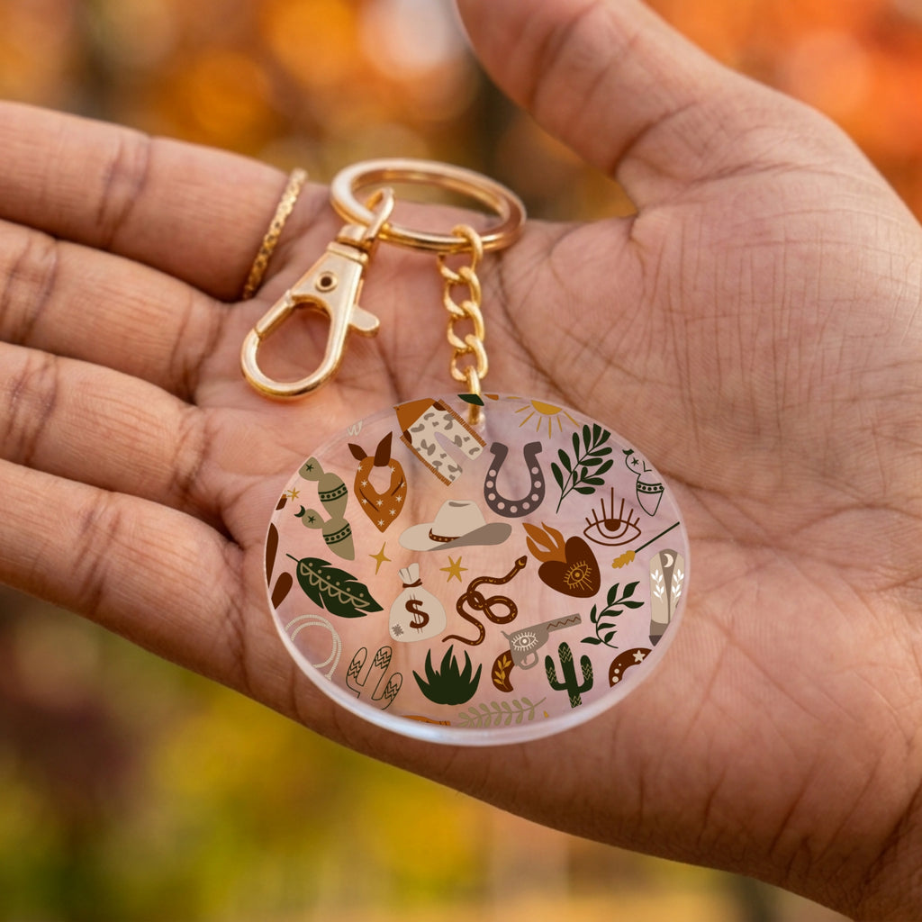 A round acrylic keychain with a gold-tone clasp and chain, held in a person's hand. The keychain features a clear circular charm decorated with various small illustrated icons including a horseshoe, cactus, snake, boots, and stars.