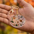 A round acrylic keychain with a gold-tone clasp and chain, held in a person's hand. The keychain features a clear circular charm decorated with various small illustrated icons including a horseshoe, cactus, snake, boots, and stars.