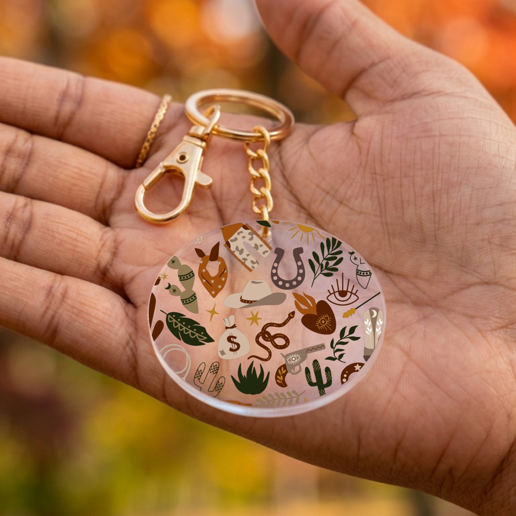 A round acrylic keychain with a gold-tone clasp and chain, held in a person's hand. The keychain features a clear circular charm decorated with various small illustrated icons including a horseshoe, cactus, snake, boots, and stars.