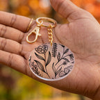 A round acrylic keychain with a black botanical line art pattern on a clear background, attached to a gold-tone chain and lobster clasp, held in a person's hand against a blurred autumn background.