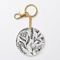 A round acrylic keychain charm featuring a black floral line art pattern over a white background with small gold dots, attached to a gold-tone split ring and lobster clasp.