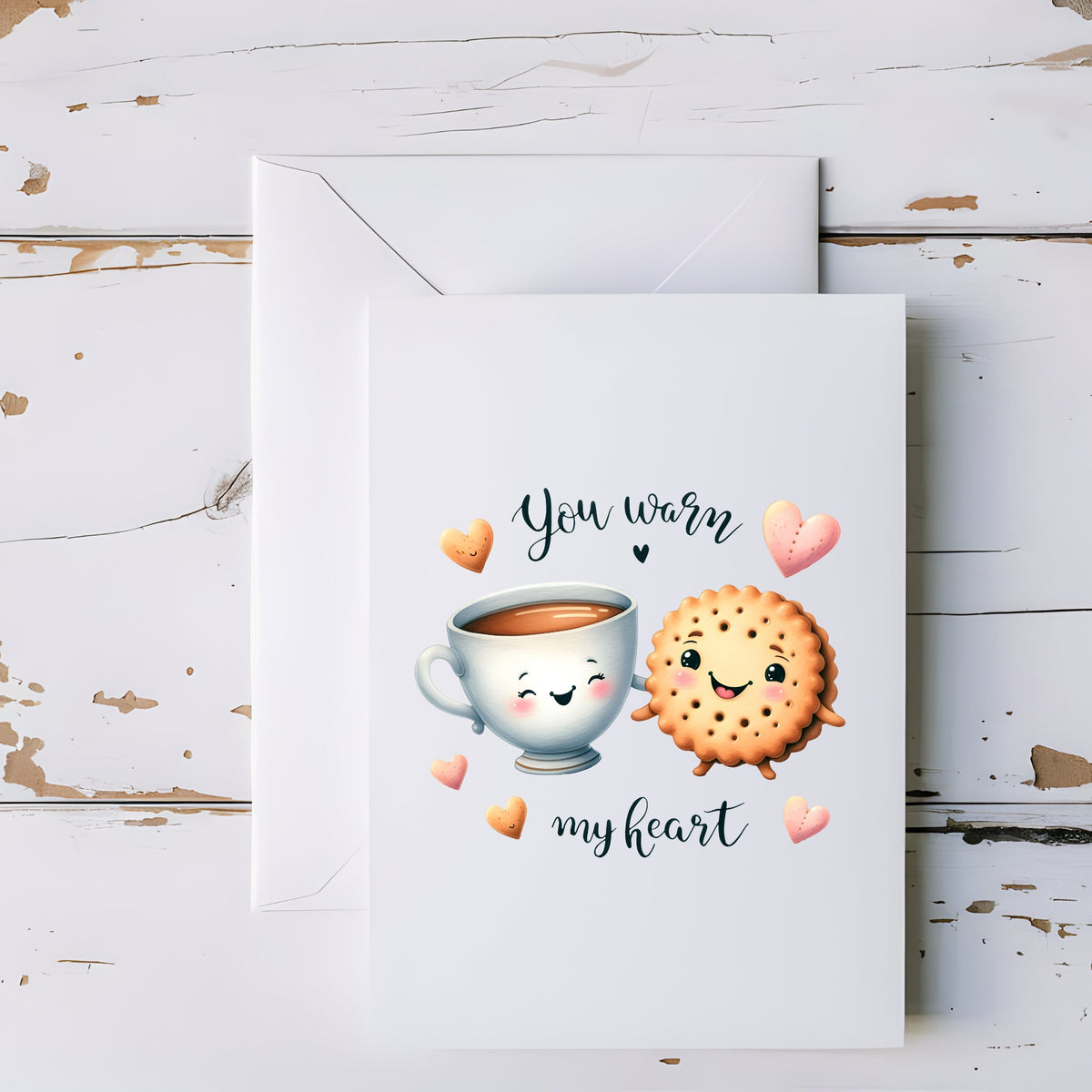 A white greeting card with a cute illustration of a smiling teacup and a smiling cookie heart, surrounded by small hearts and the words 'You Warm My Heart,' shown with a matching white envelope on a distressed white wood background.