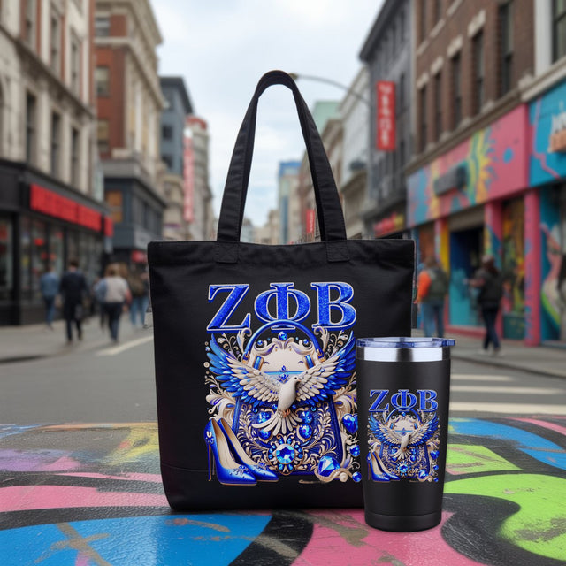 A black tote bag and matching black tumbler with a blue and white ornate crest and the Greek letters “ZΦB” on a blurred city street background.