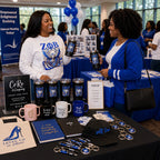 Two women standing at a promotional booth with branded tumblers, pens, notebooks, and bags in a bright indoor event space. The table displays blue-and-white themed merchandise, including a black cap with 'self love' text, keychains, pens, notebooks, and mugs.