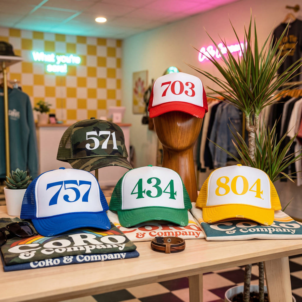 Five trucker hats with area code numbers and CoRo & Company shirts on a table in a store setting.