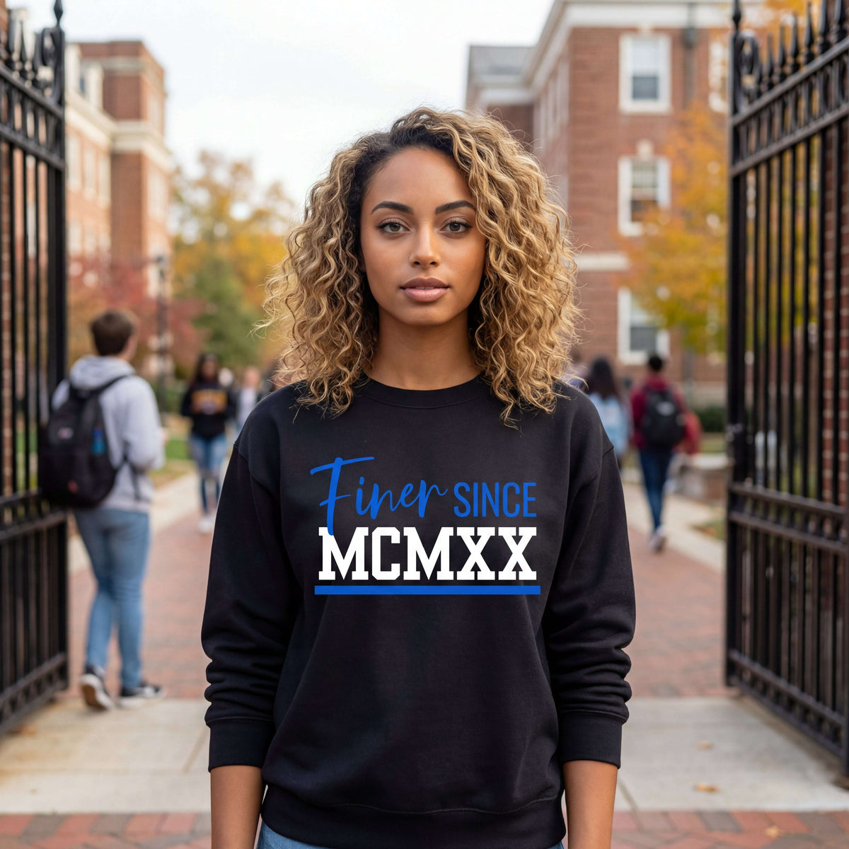 A Zeta woman wearing a black Finer Since MCMXX sweatshirt, standing in an outdoor setting with people and buildings in the background.