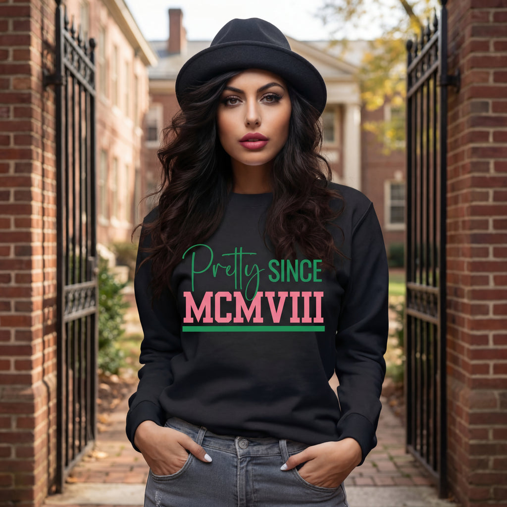 An AKA Woman wearing a black sweatshirt with 'Pretty Since MCMVIII' text, standing in front of a college building.