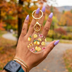A hand with lavender nail polish holds a round clear acrylic keychain featuring yellow lemon illustrations and small pink bow details, with gold-tone metal rings and clasp, against a blurred autumn park background.