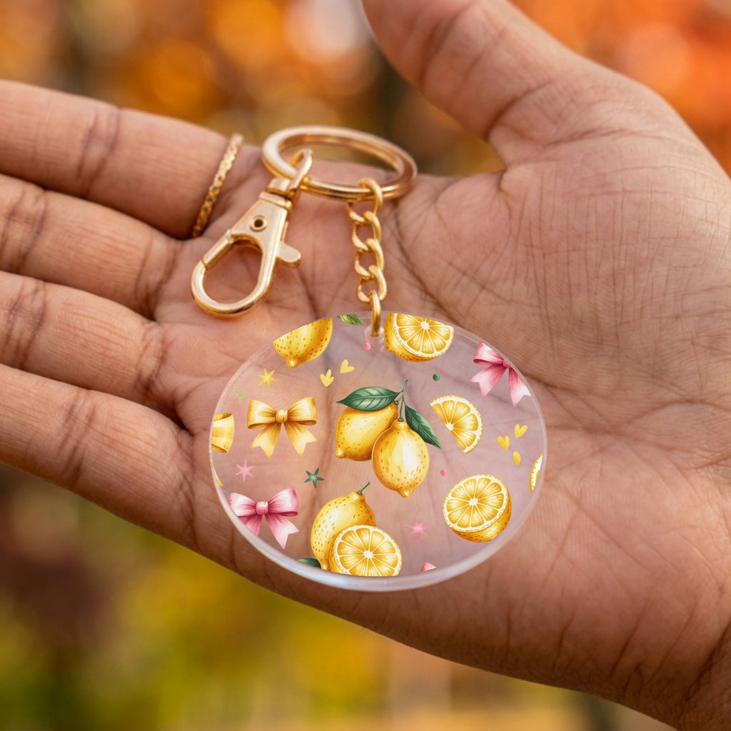 A hand holding a round acrylic keychain charm decorated with yellow lemons, pink bows, and small gold stars on a translucent background, with a gold clasp and chain.