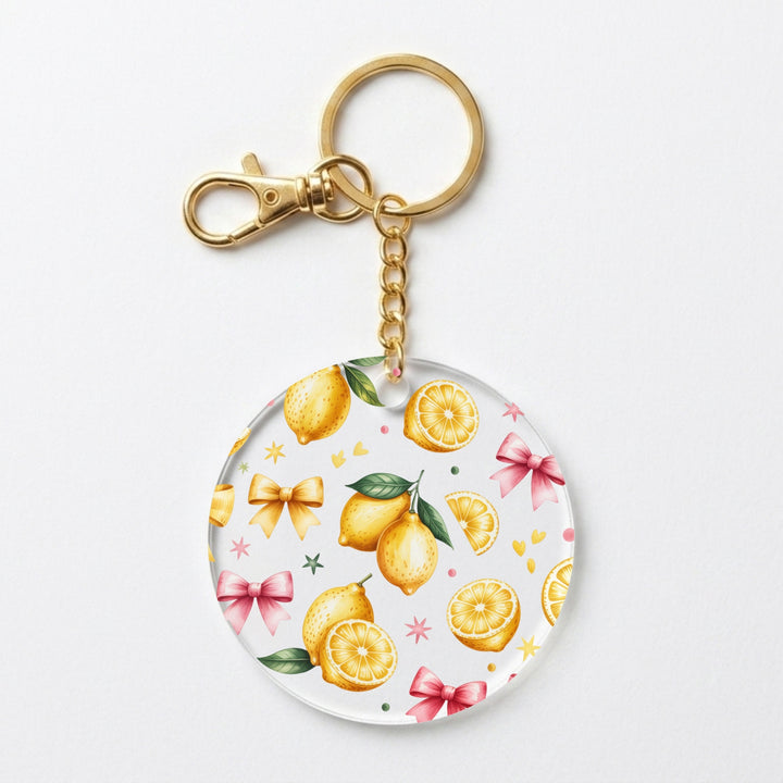 A round acrylic keychain featuring a watercolor lemon and pink bow pattern on a white background, attached to a gold-tone metal split ring and clasp.