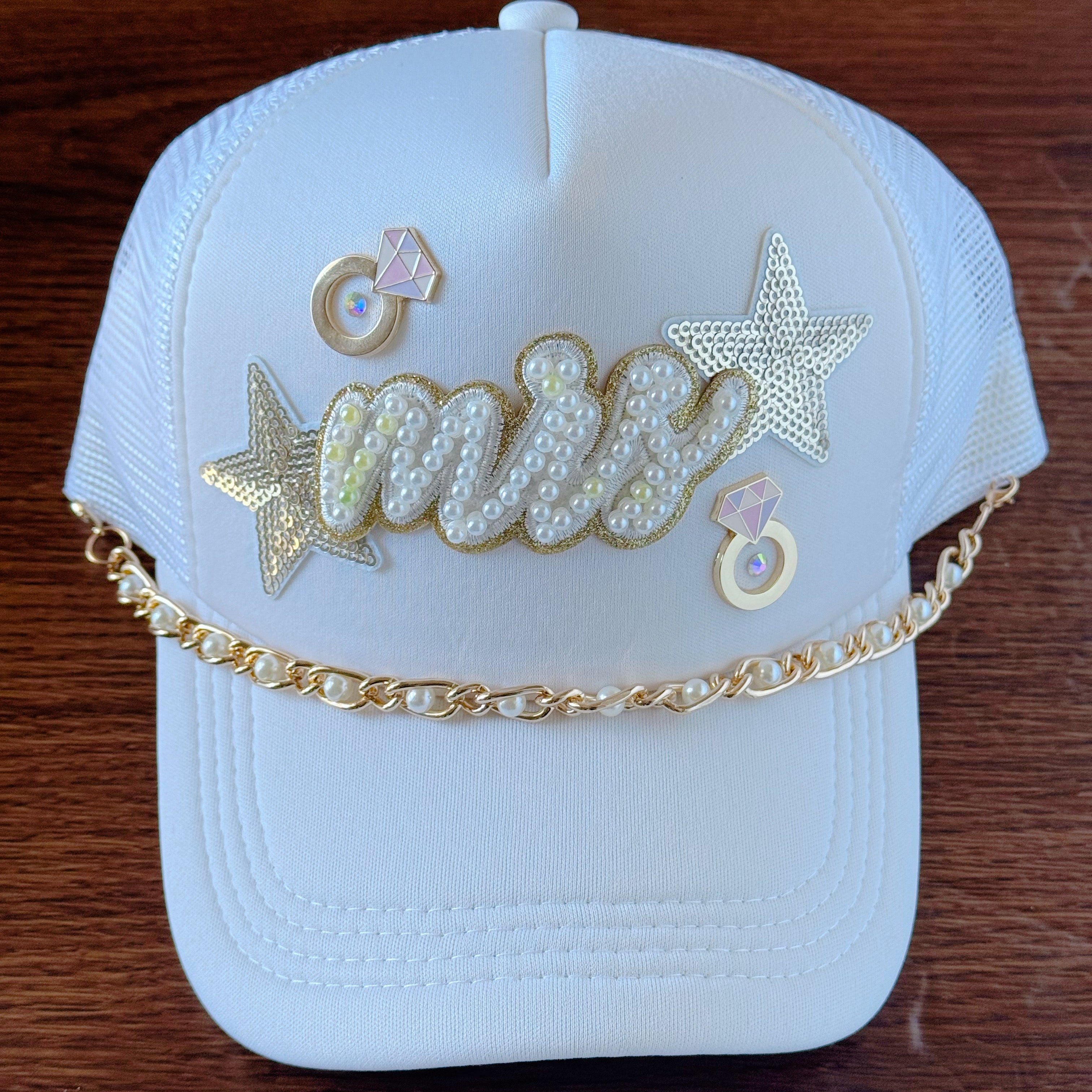 White bridal trucker cap with "Mrs" patch decorated in white and yellow pearls, gold pearl hat chain, 2 engagement ring pins, and stars on a wooden surface