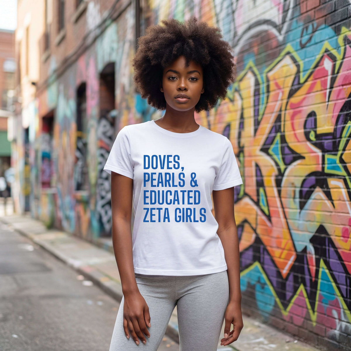 A Zeta wearing a white Doves, Pearls, & Educated Zeta Girls t-shirt with blue text in an urban setting with colorful graffiti.