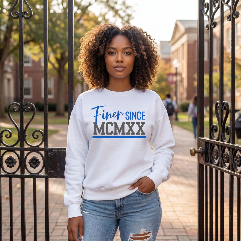 A Zeta woman wearing a white Finer Since MCMXX sweatshirt, standing in front of an iron gate of a college campus.