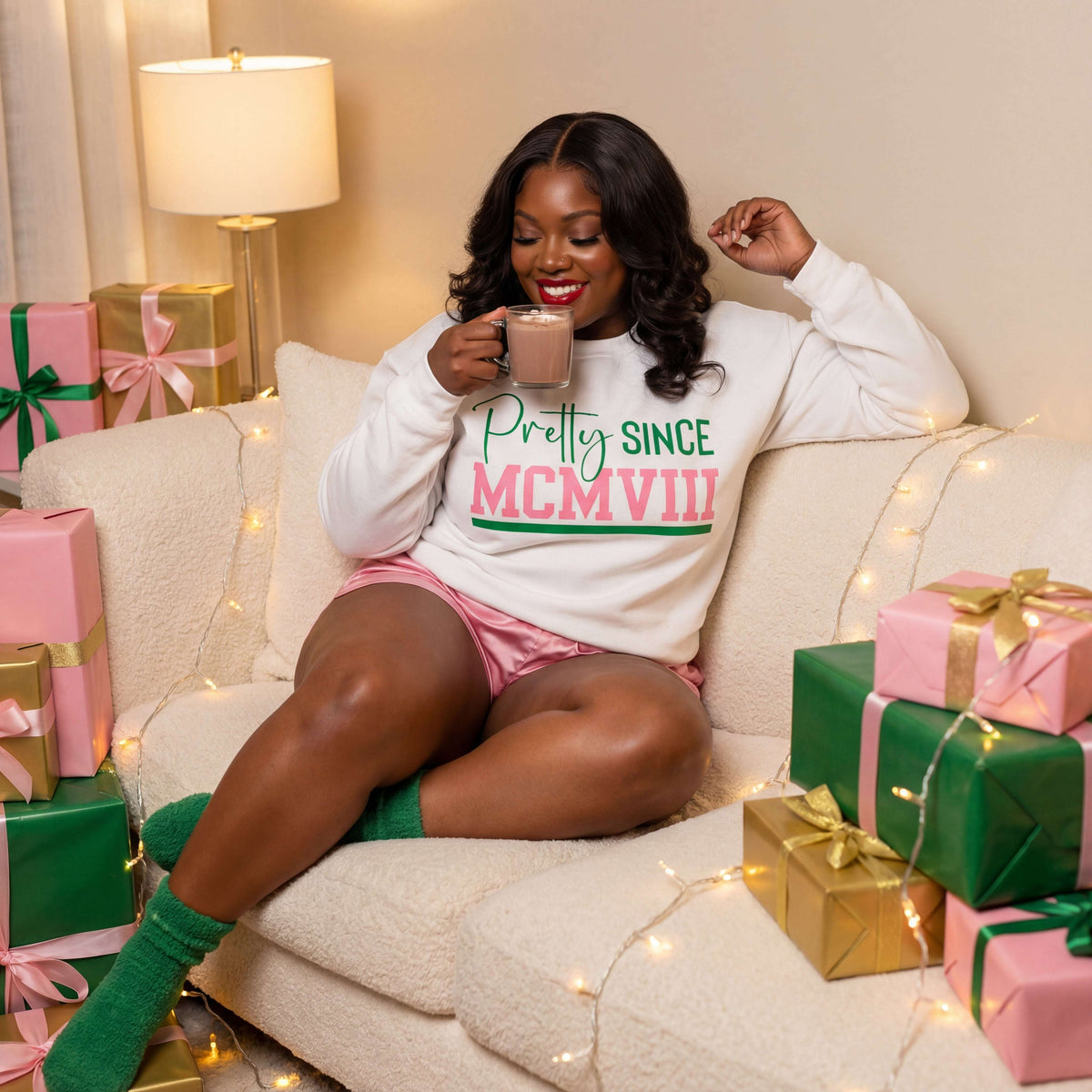 An AKA Woman sitting on a couch with Christmas presents and drinking coffee wearing a white Pretty Since MCMVIII sweatshirt.